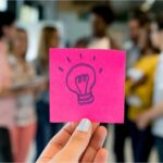 A hand holding a pink post it note with a light bulb drawn on it.